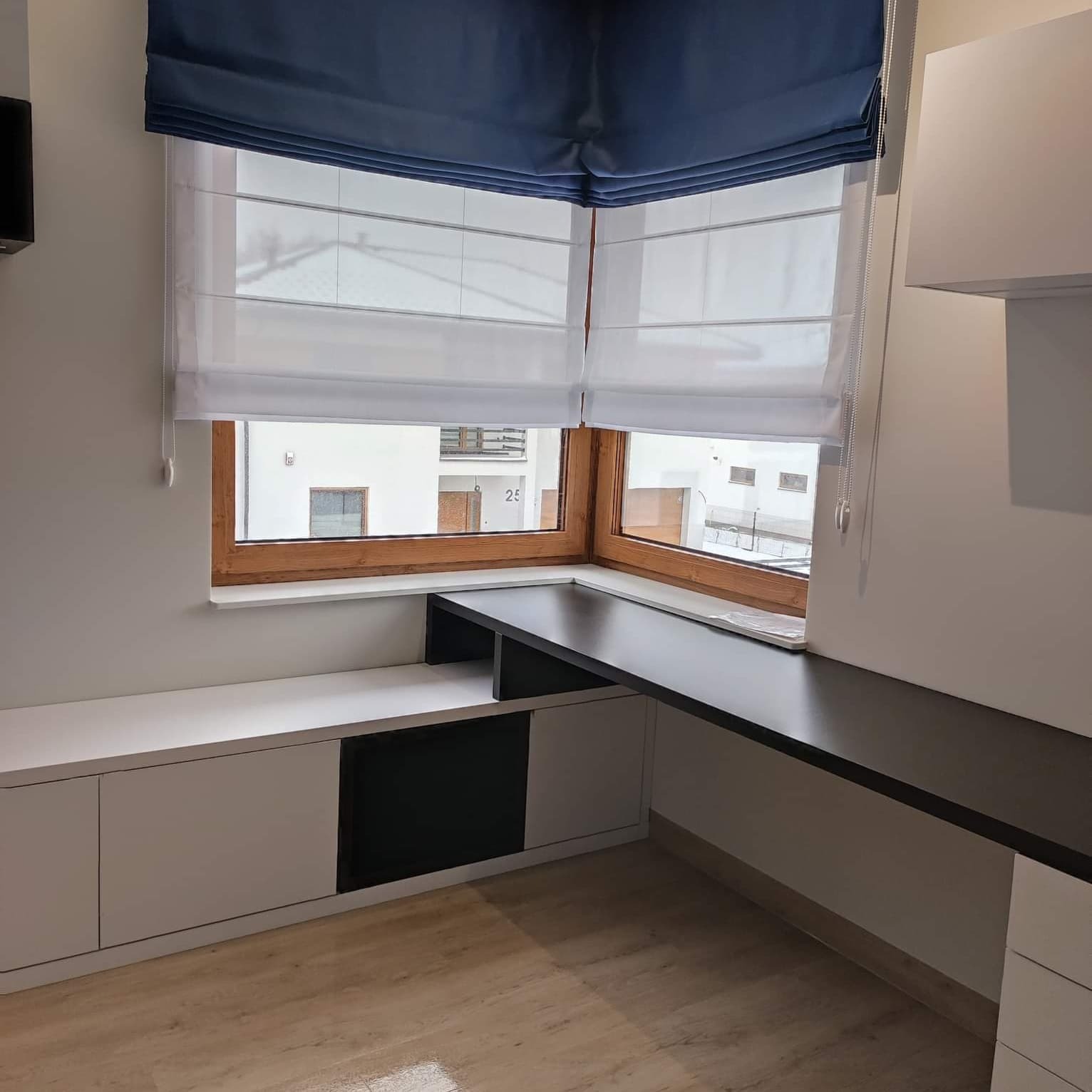 Biała roleta rzymska z granatowym pasem u góry, zamontowana w oknie nad biurkiem w nowoczesnym pokoju.