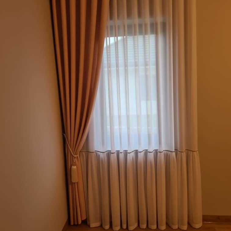 Zasłony w odcieniach beżu i bieli przy oknie, z dekoracyjnym wiązaniem i frędzlem po lewej stronie, delikatna falbana u dołu.