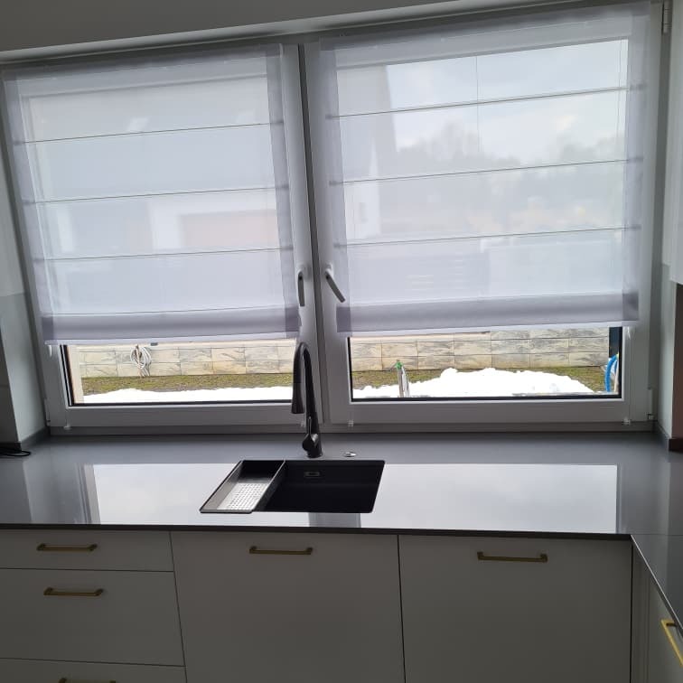 Białe rolety rzymskie na oknach nad szarym blatem kuchennym z czarnym zlewem, widok na ośnieżony ogród.
