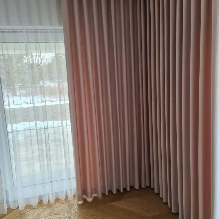 Eleganckie, długie zasłony w odcieniach pudrowego różu i beżu, z widocznym marszczeniem, zawieszone obok okna z delikatną, białą firanką, na tle drewnianej podłogi i zimowego krajobrazu za oknem.