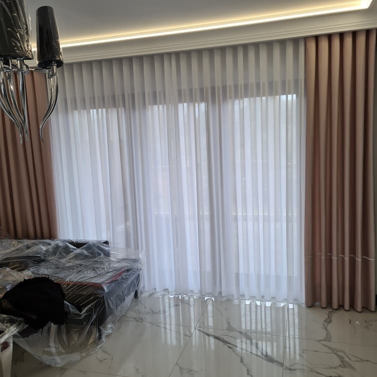 Eleganckie, dwuwarstwowe zasłony: transparentna, biała firana w połączeniu z beżowymi zasłonami zaciemniającymi, widoczne na tle marmurowej podłogi i częściowo zakrytego mebla folią ochronną.