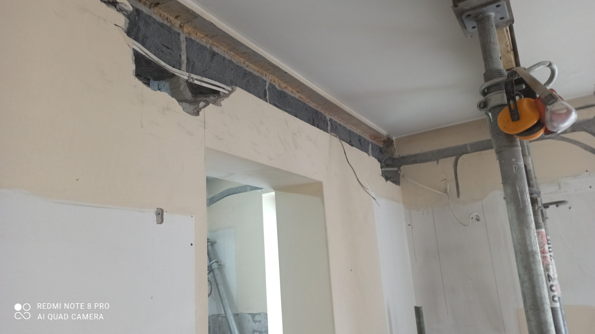 Fragment ściany z wyciętym otworem, widoczne przewody elektryczne i fragment stropu, podpora budowlana z żółtymi słuchawkami ochronnymi w tle.