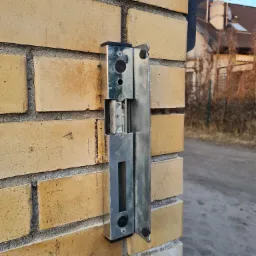 Elektrozaczep domofonu zamontowany na murze z cegły klinkierowej, widoczny zamek i elementy mocujące.