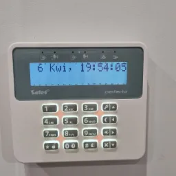 Panel sterowania alarmu Satel Perfecta z podświetlanymi przyciskami, wyświetlający datę 6 Kwi i godzinę 19:54:05, zamontowany na jasnej ścianie.