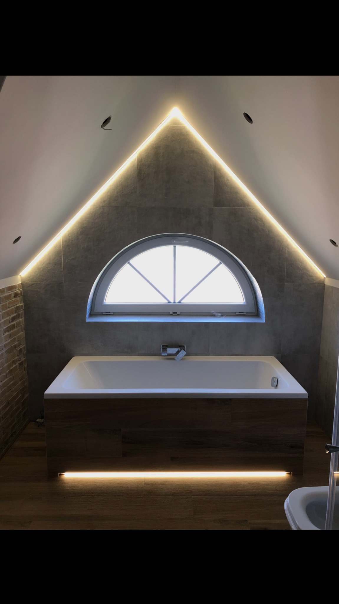 Nowoczesna łazienka na poddaszu z wanną obudowaną drewnem, oświetleniem LED wzdłuż krawędzi dachu i pod wanną, oraz półokrągłym oknem.