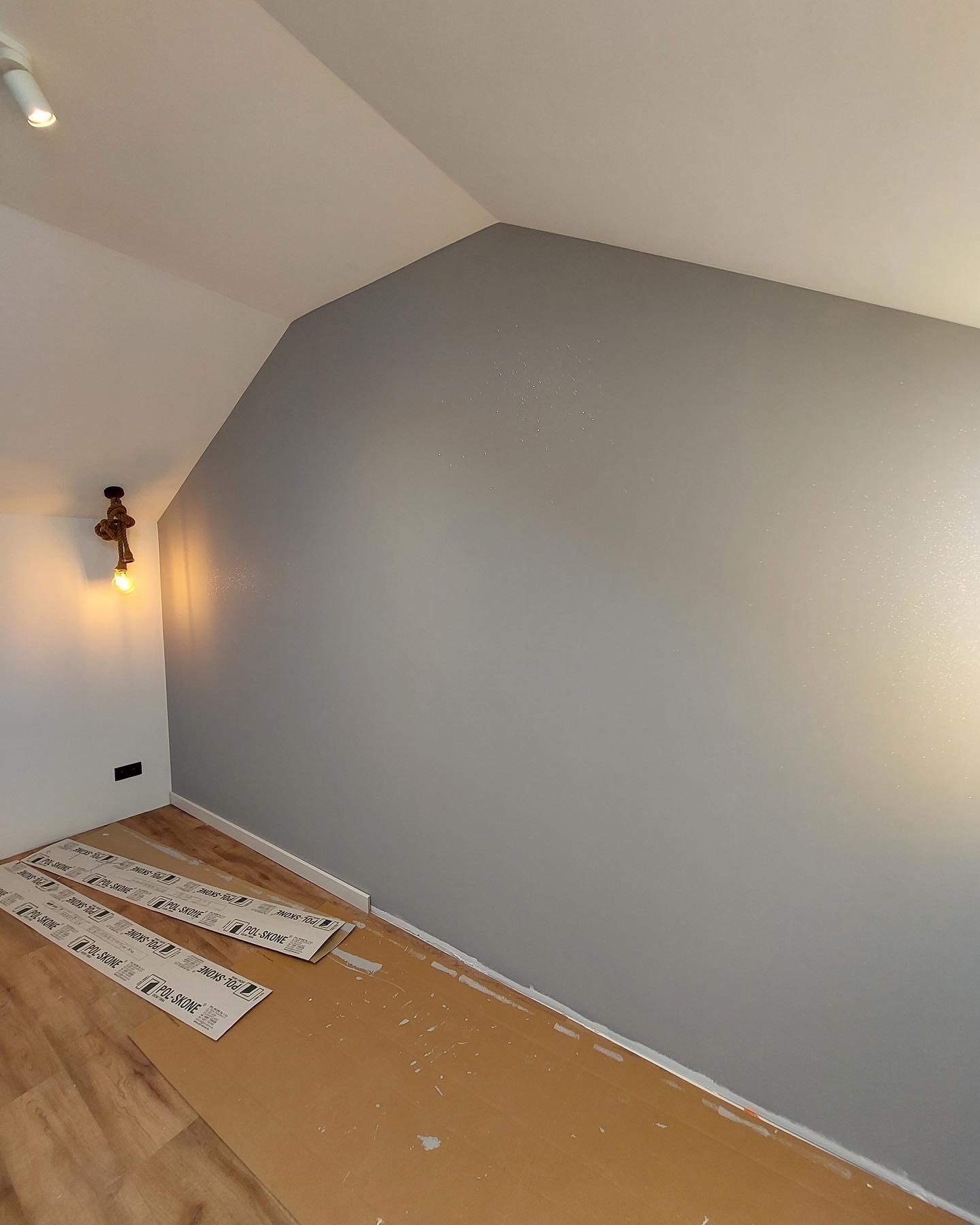 Świeżo pomalowana szara ściana w pokoju na poddaszu, z widocznymi zabezpieczeniami podłogi i lampą ze sznura w rogu, sugerująca etap wykończeniowy prac.