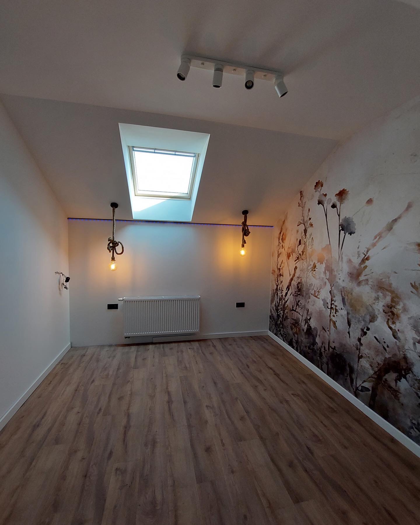 Poddasze po remoncie z drewnianą podłogą, grzejnikiem, lampami wiszącymi na sznurze i ścianą z motywem kwiatowym, oświetlone światłem z okna dachowego.