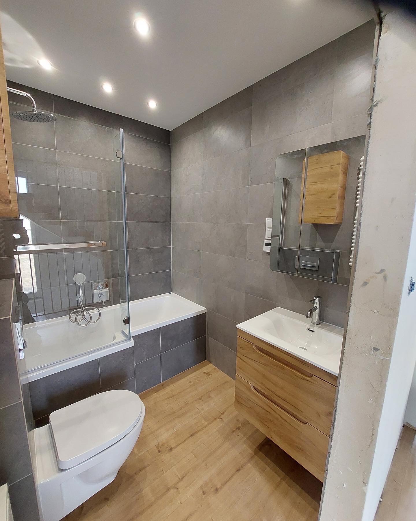 Nowoczesna łazienka z szarymi płytkami, drewnianą szafką pod umywalką i kabiną nawannową, widok na fragment nieukończonej ściany po prawej.