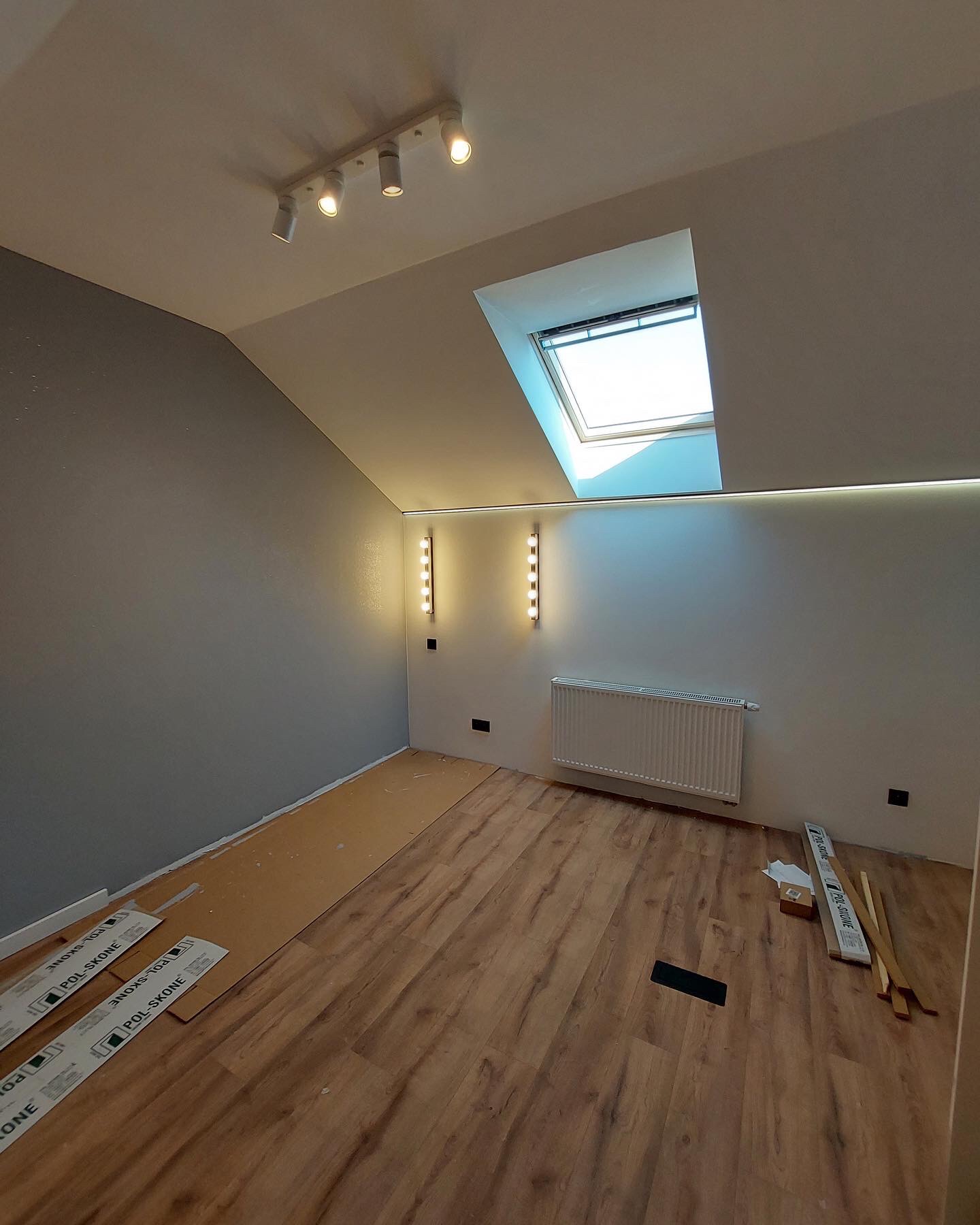 Poddasze po częściowym położeniu paneli podłogowych, widoczne paczki paneli przy ścianach, grzejnik, okno dachowe i nowoczesne oświetlenie.