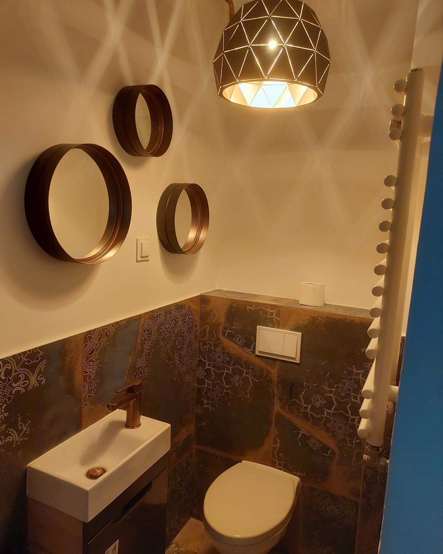 Nowoczesna łazienka z okrągłymi lustrami, geometryczną lampą wiszącą i dekoracyjnymi płytkami ściennymi w odcieniach brązu i złota, z umywalką nablatową i armaturą w kolorze miedzi.