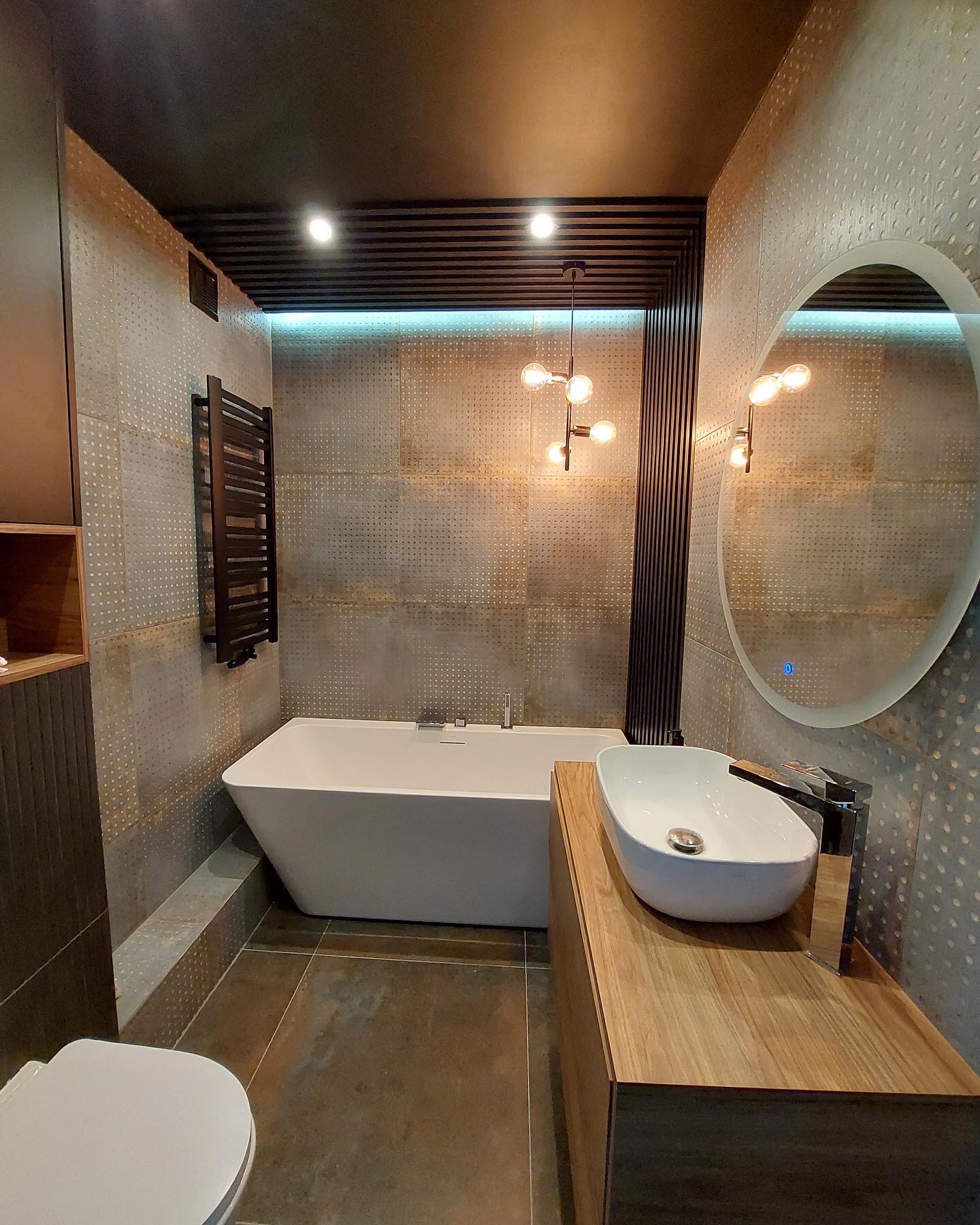 Nowoczesna łazienka z wanną, umywalką nablatową na drewnianej szafce, okrągłym lustrem z podświetleniem LED i dekoracyjnymi płytkami ściennymi z efektem rdzy.