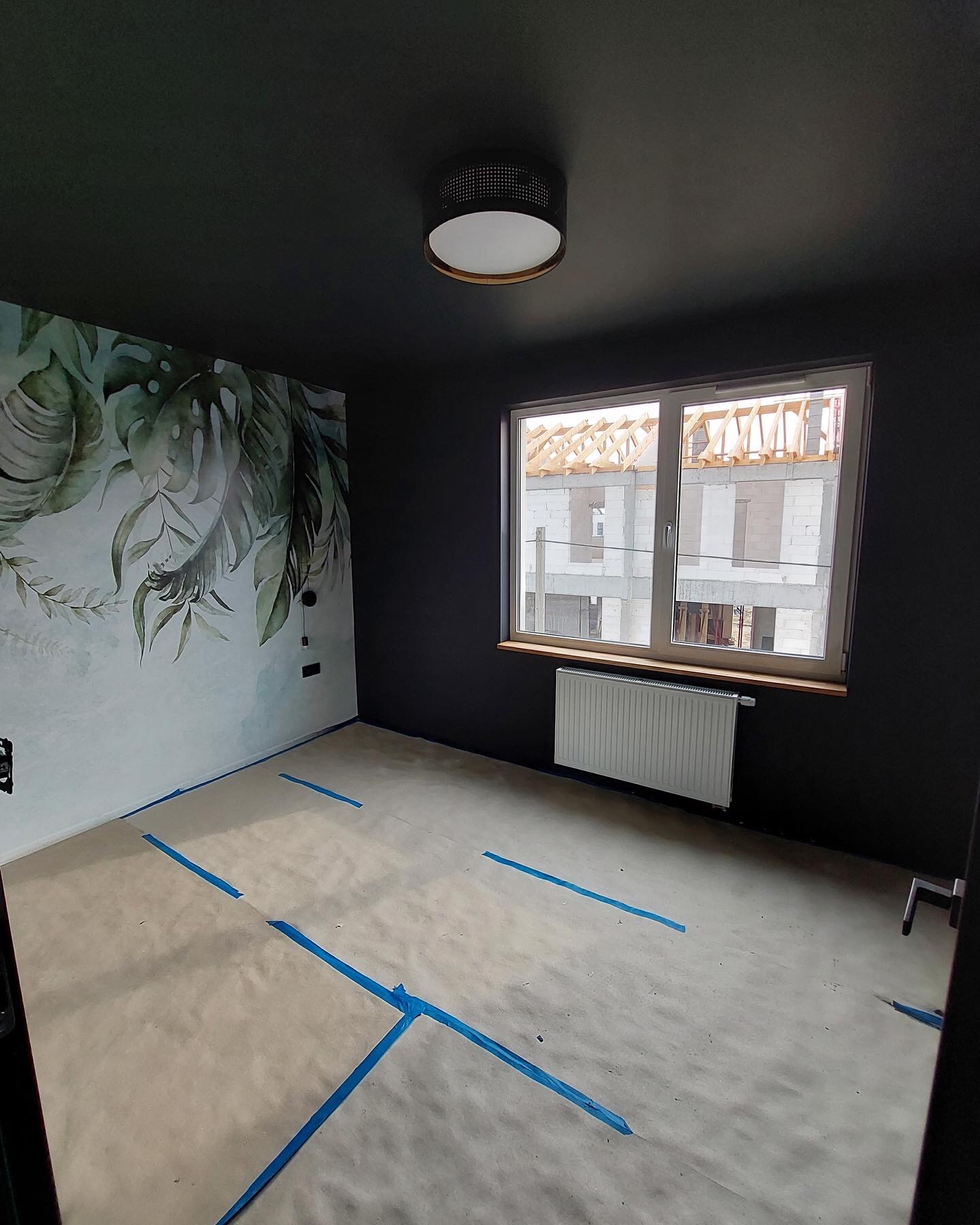 Pokój z ciemnymi ścianami i fototapetą z motywem liści; podłoga zabezpieczona papierem i niebieską taśmą malarską, widok przez okno na budowę.