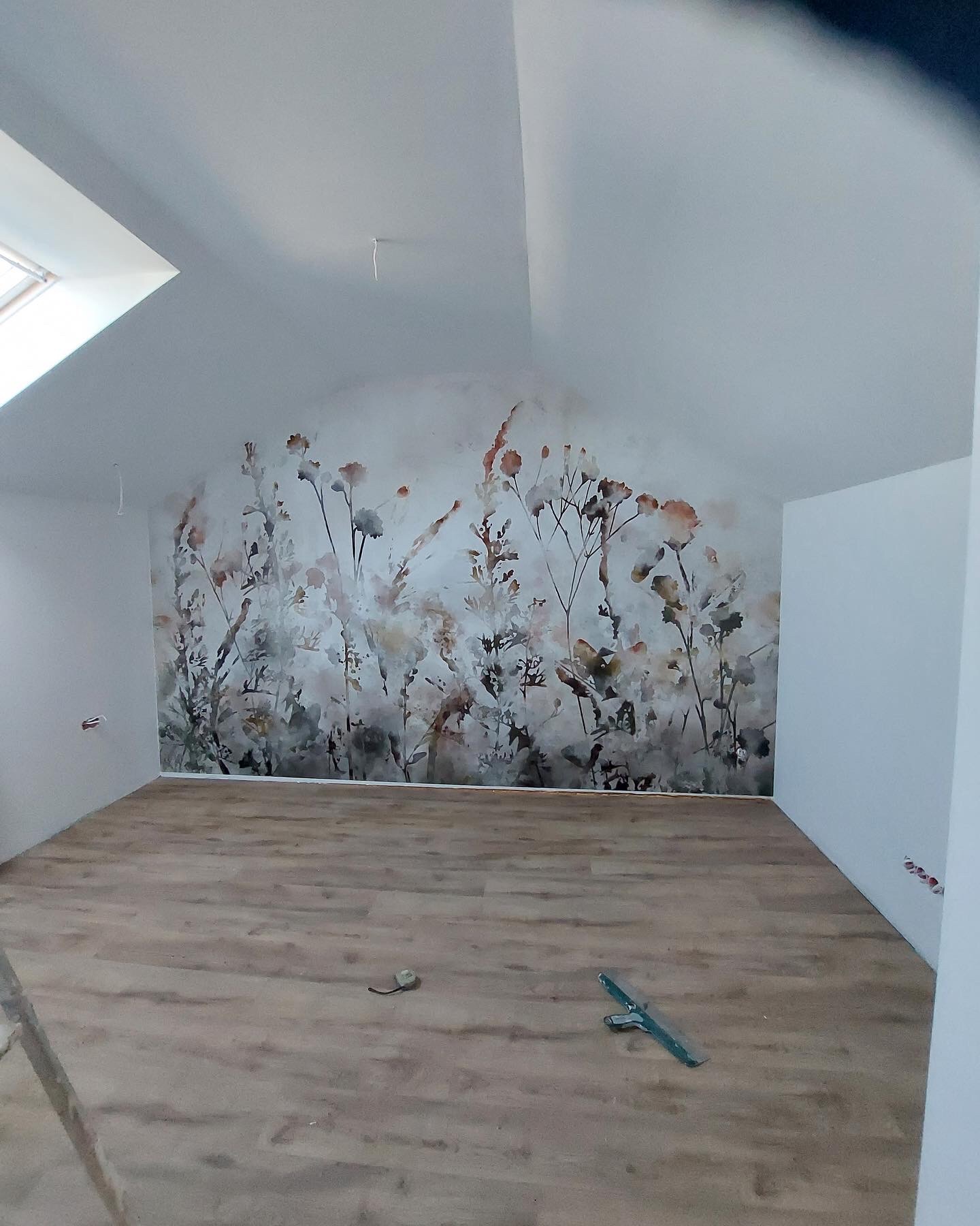 Pokój na poddaszu po remoncie z nową fototapetą z motywem łąki i ułożonymi panelami podłogowymi; widoczne narzędzia na podłodze i okno dachowe.