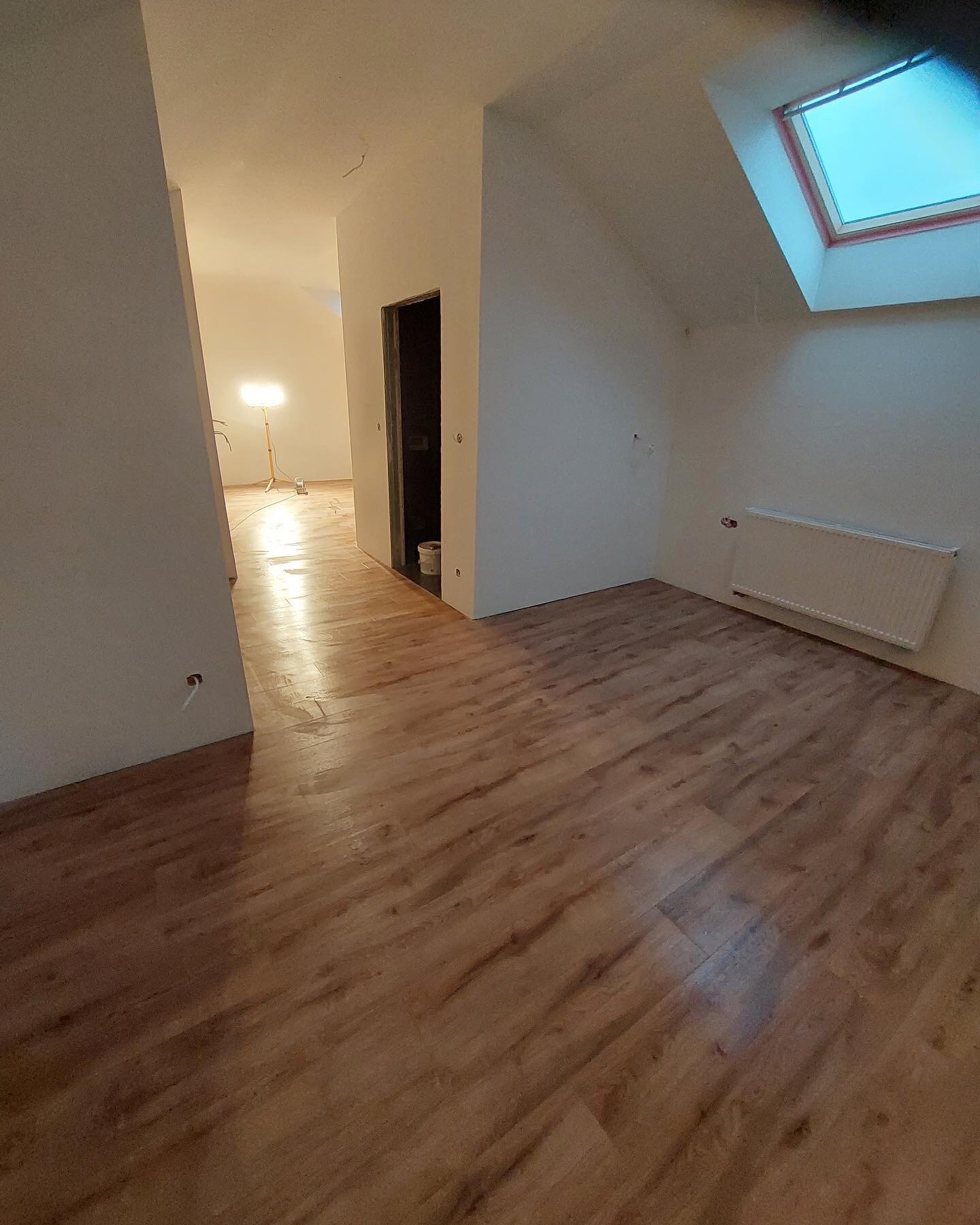 Poddasze z nowo ułożonymi panelami podłogowymi w odcieniach brązu, świeżo pomalowanymi ścianami na biało, widocznym grzejnikiem i oknem dachowym z niebieskim niebem w tle; lampa budowlana w tle...