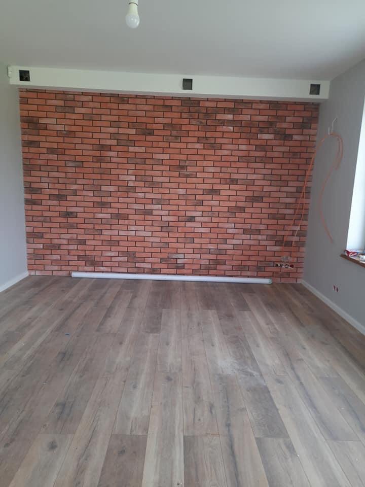 Pokój po remoncie z ceglaną ścianą i panelami podłogowymi, widoczne otwory wentylacyjne pod sufitem i tymczasowe oświetlenie.