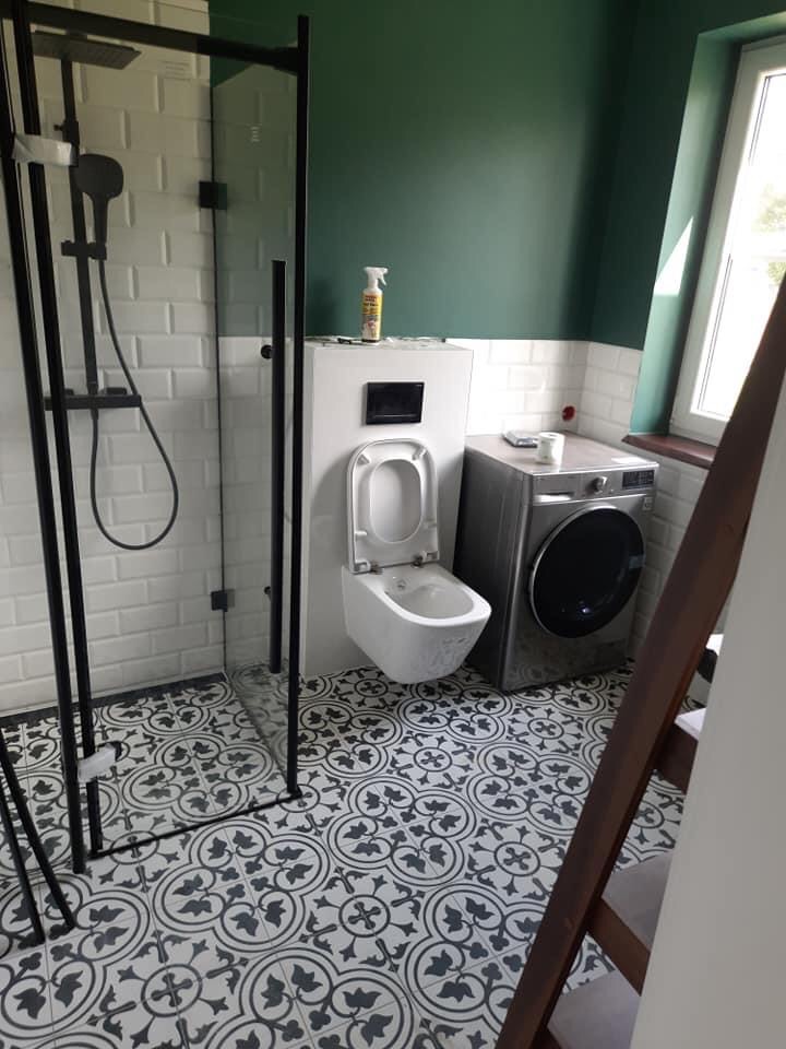 Nowoczesna łazienka z czarną kabiną prysznicową, wiszącą toaletą i pralką, wykończona białymi płytkami typu metro i wzorzystą podłogą w odcieniach szarości, z zieloną ścianą w tle.