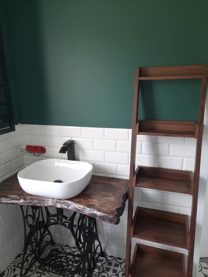 Biała umywalka nablatowa na blacie z surowego drewna osadzonym na żeliwnej podstawie starej maszyny do szycia, białe kafelki typu metro i ciemnozielona ściana w tle, obok drewniany regał drabinkowy.