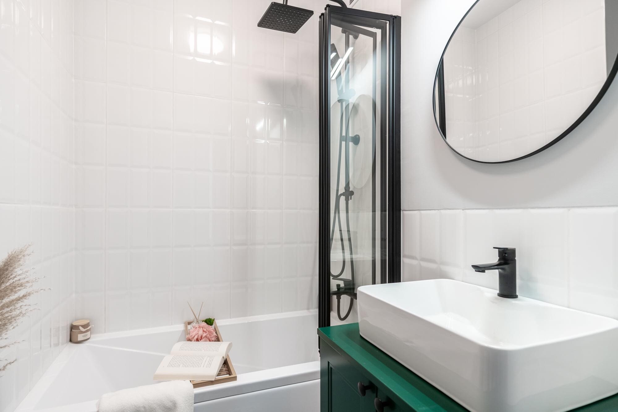 Biała łazienka z wanną, kabiną prysznicową z czarną ramą, umywalką nablatową na zielonej szafce i okrągłym lustrem, dekorowana książką i ozdobami.