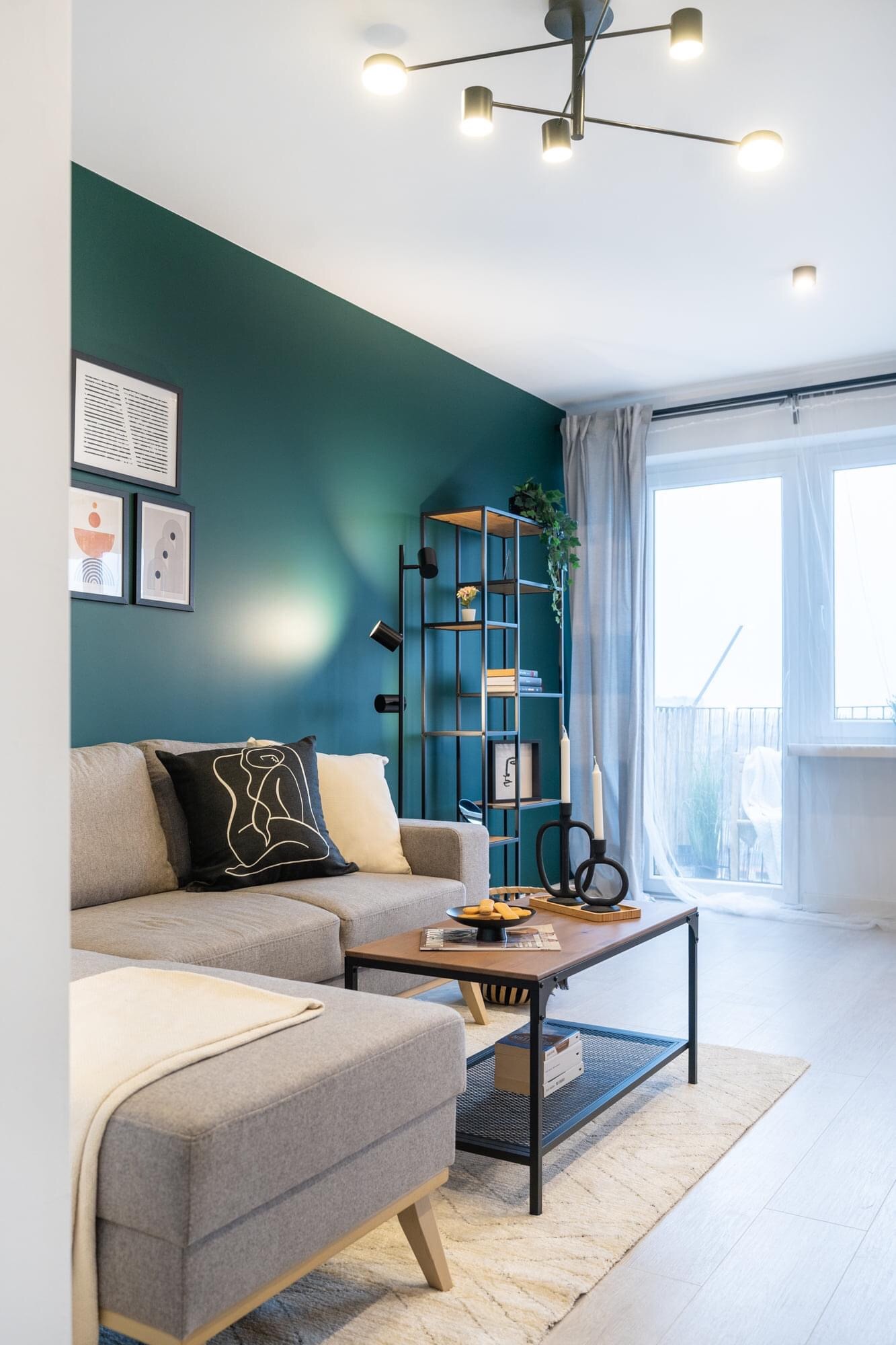 Nowoczesny salon z turkusową ścianą, szarą sofą, drewnianym stolikiem kawowym i metalowym regałem, oświetlony lampą sufitową w stylu industrialnym.