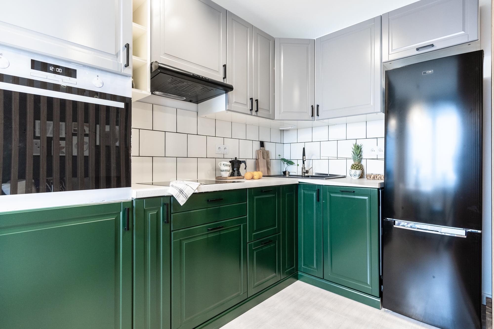 Nowoczesna kuchnia z zielonymi szafkami dolnymi i szarymi górnymi, czarny piekarnik i lodówka, białe płytki na ścianie.