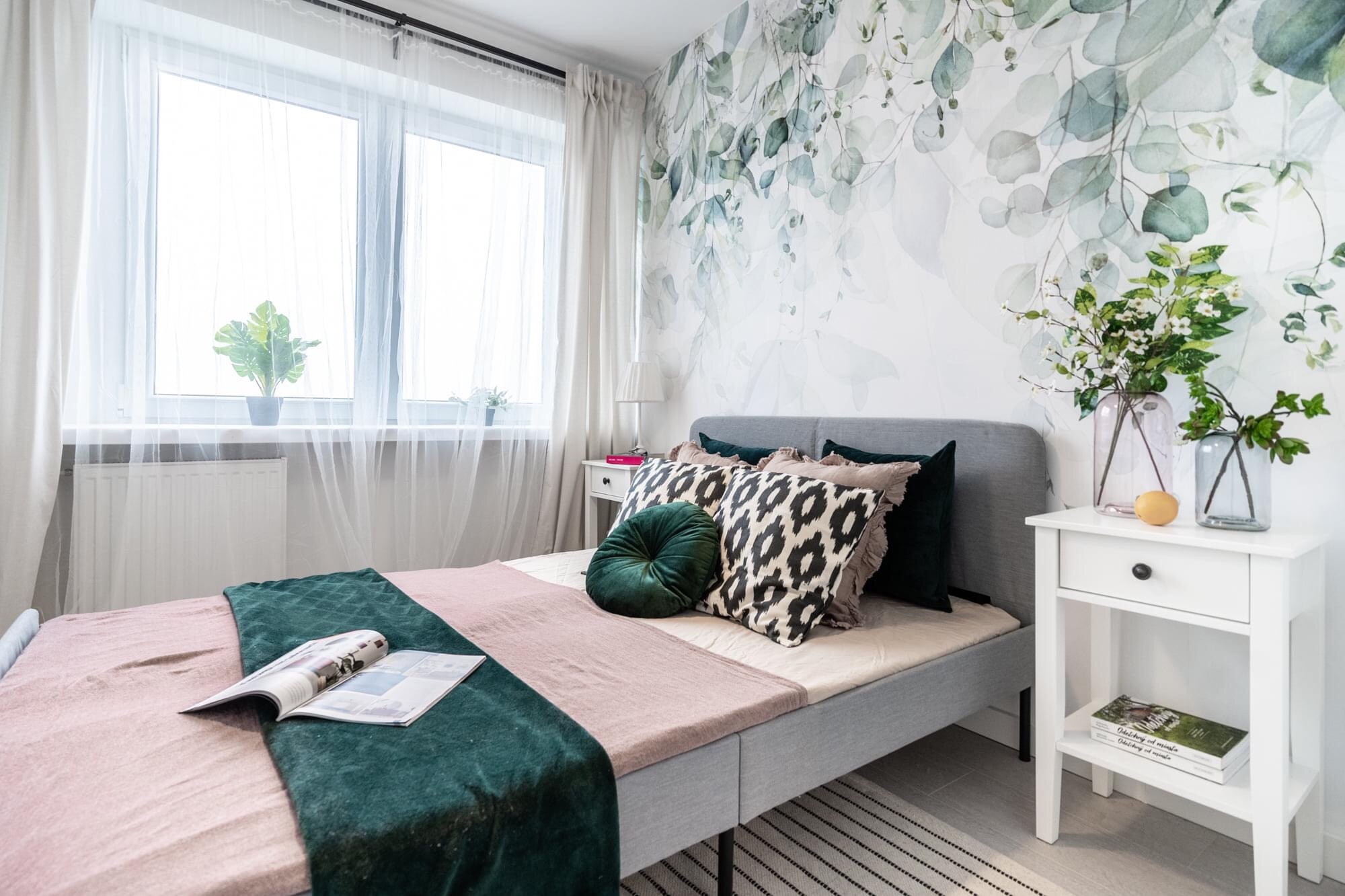 Jasna sypialnia z łóżkiem, wzorzystą tapetą w liście i kwiatami w wazonie na szafce nocnej. Na łóżku leży otwarty magazyn wnętrzarski.
