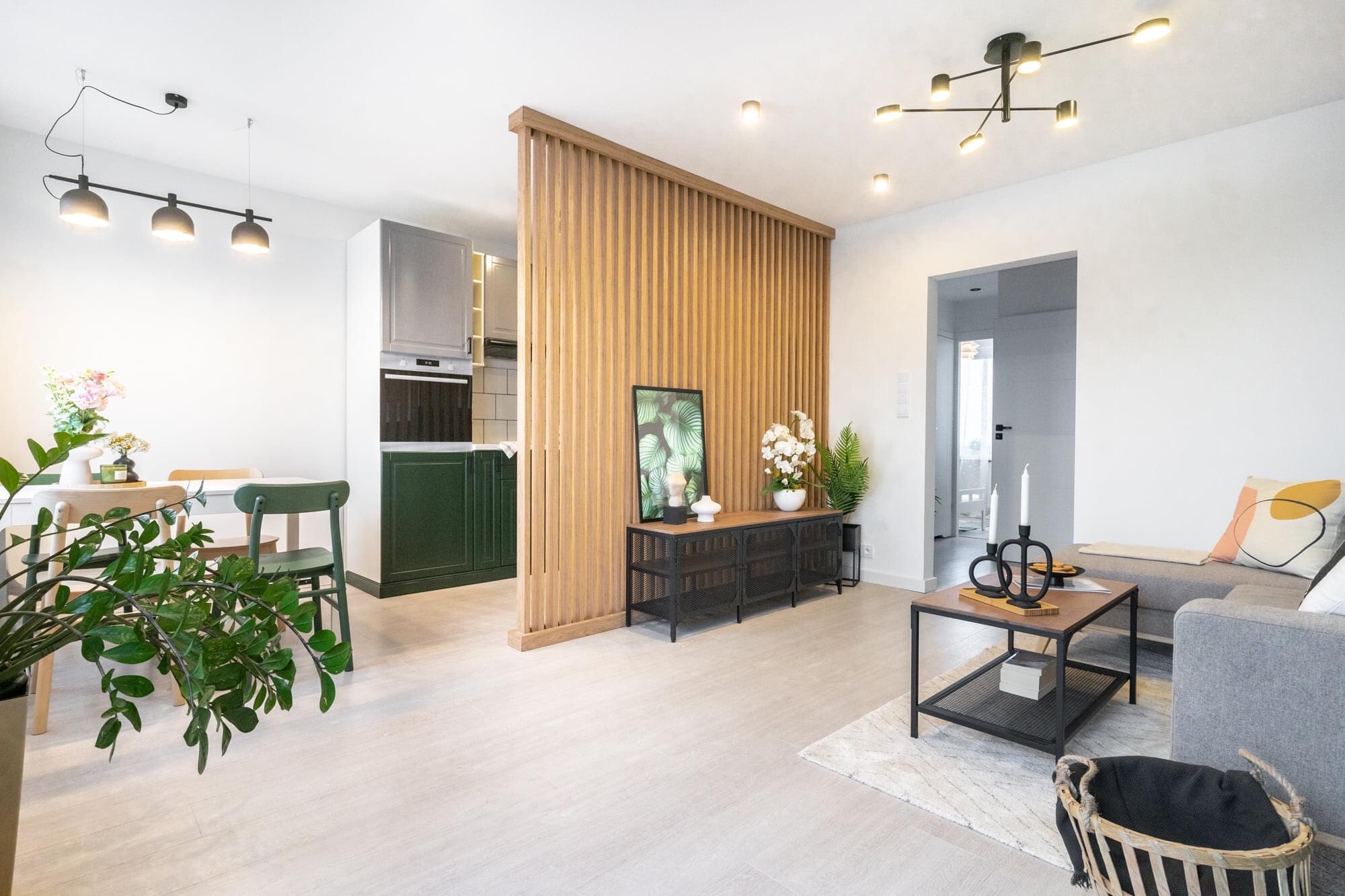 Jasne wnętrze mieszkania z drewnianą ażurową ścianką działową, zielonymi akcentami w kuchni i roślinami doniczkowymi, ukazujące aranżację po remoncie.