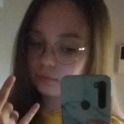 Młoda osoba w okularach robi selfie, pokazując gest 'metal' jedną ręką, ubrana w żółtą bluzę z napisem.