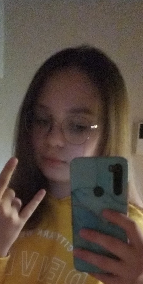 Młoda osoba w okularach robi selfie, pokazując gest 'metal' jedną ręką, ubrana w żółtą bluzę z napisem.