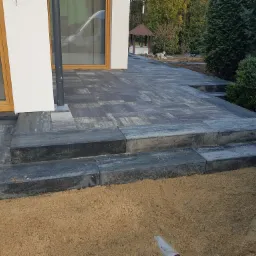 Ułożona kostka brukowa w odcieniach szarości tworząca taras i schody przy budynku z białymi ścianami i drewnianymi oknami; widoczny piasek podsypkowy, w tle altana ogrodowa