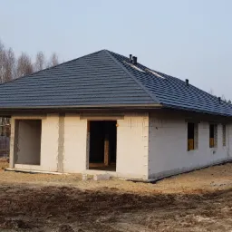 Surowy stan domu z białego bloczka, z ukończonym dachem z ciemnej blachy, widok z zewnątrz, w tle drzewa, na pierwszym planie ziemia.