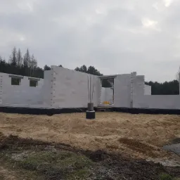 Wznoszone ściany z białych bloczków na etapie budowy domu, widoczne zbrojenia, izolacja fundamentów i piaszczyste podłoże, fragment białego samochodu dostawczego z prawej strony kadru.