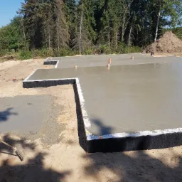 Wylany fundament pod dom z widoczną izolacją pionową i wystającymi rurami, w tle drzewa i usypana ziemia.