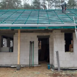 Budowa domu w stanie surowym otwartym: ściany z białego bloczka, częściowo ułożone zielone dachówki, drabina oparta o dach, pracownik na dachu, widoczne podpory budowlane i narzędzia.