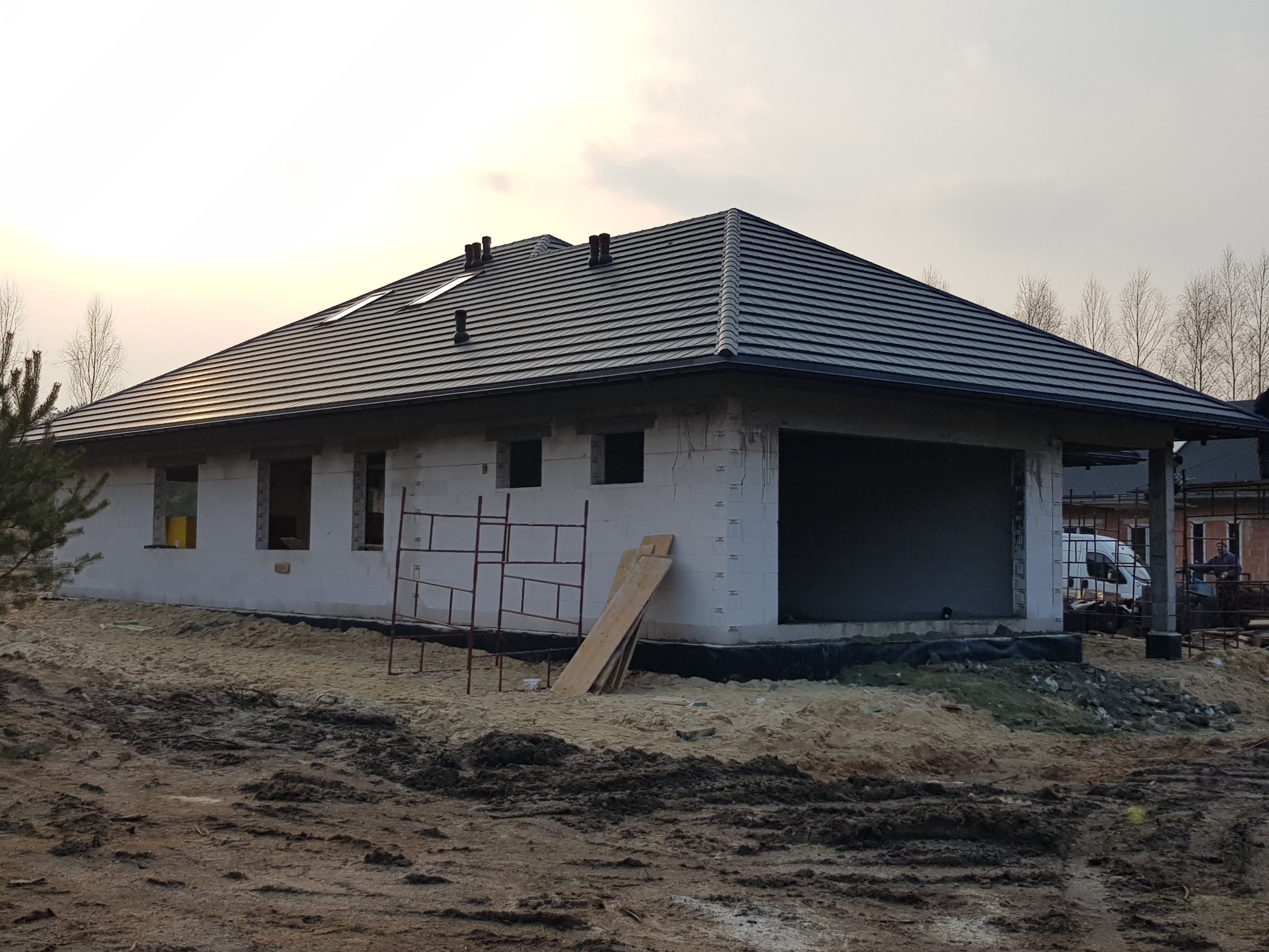 Surowy stan domu jednorodzinnego z białego bloczka, z ciemnoszarą dachówką, widoczne rusztowanie i deski, teren wokół domu w stanie budowy.