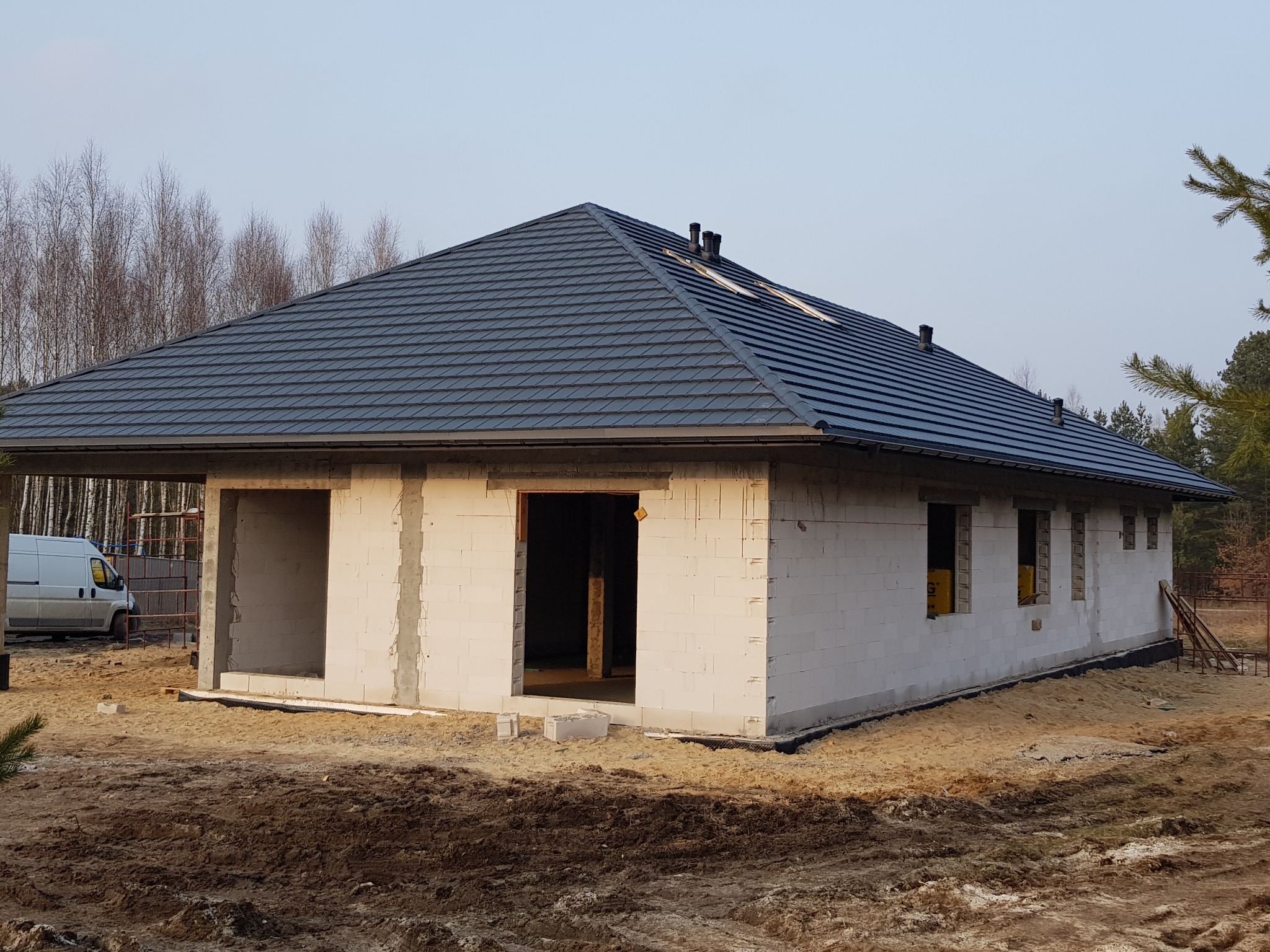 Surowy stan domu z białego bloczka, z ukończonym dachem z ciemnej blachy, widok z zewnątrz, w tle drzewa, na pierwszym planie ziemia.