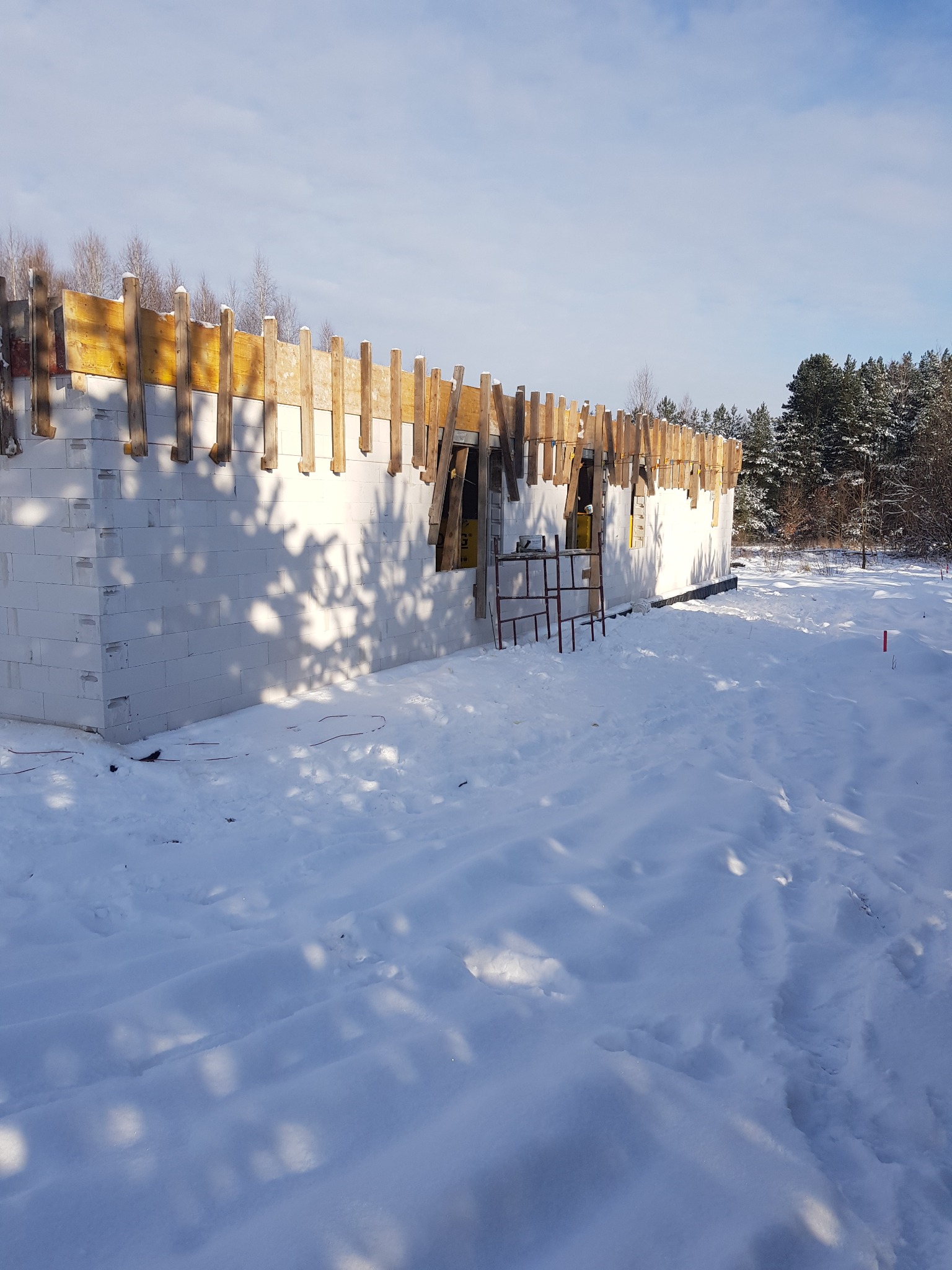 Budowa domu murowanego w zimowej scenerii: ściany z białych bloczków, drewniane szalunki pod wieniec, zasypany śniegiem teren wokół budynku, widoczny las w tle.