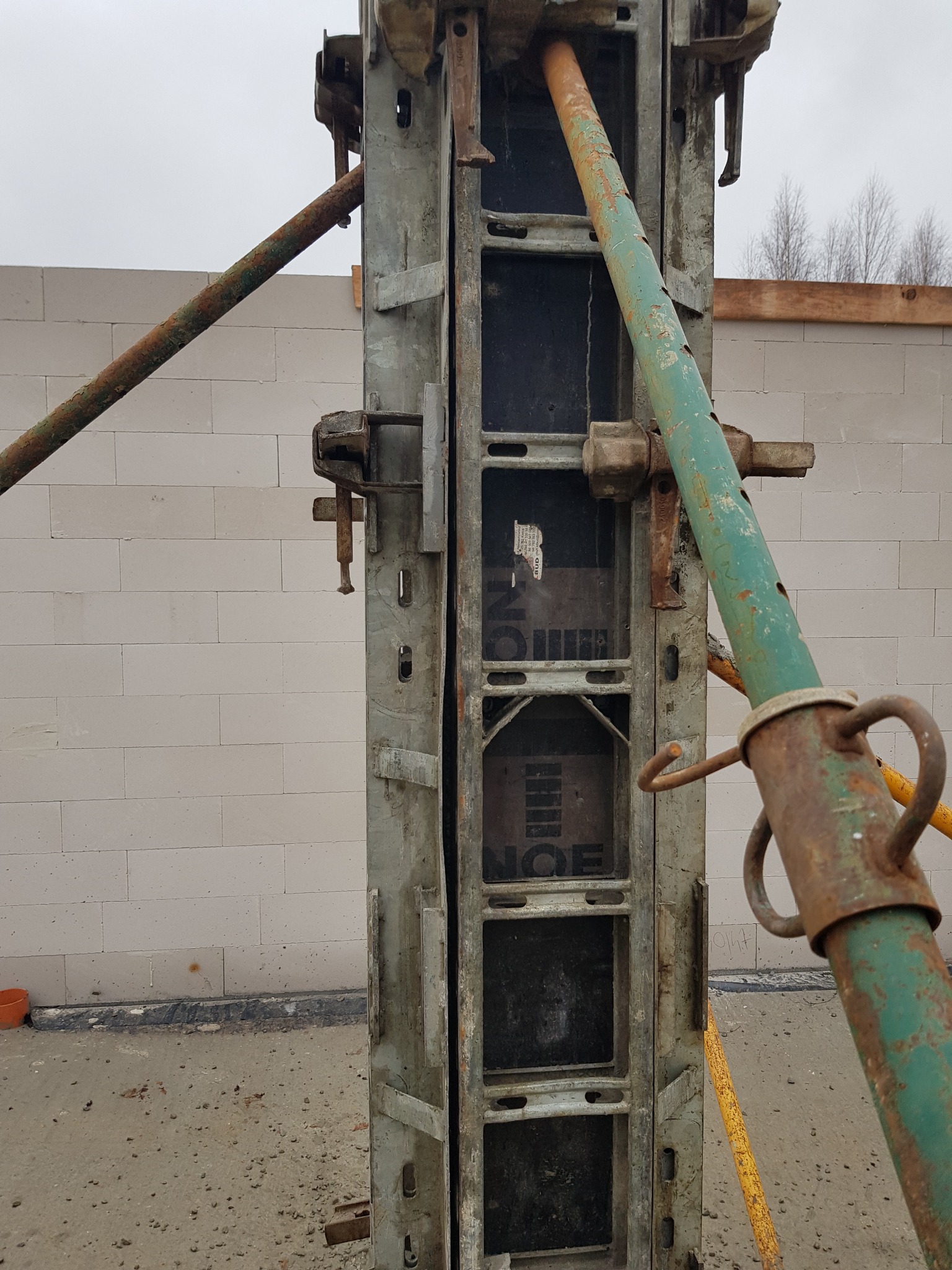 Szare szalunki kolumnowe, podparte zielonymi i żółtymi stemplami, na tle ściany z białych bloczków betonu komórkowego i szarego nieba.