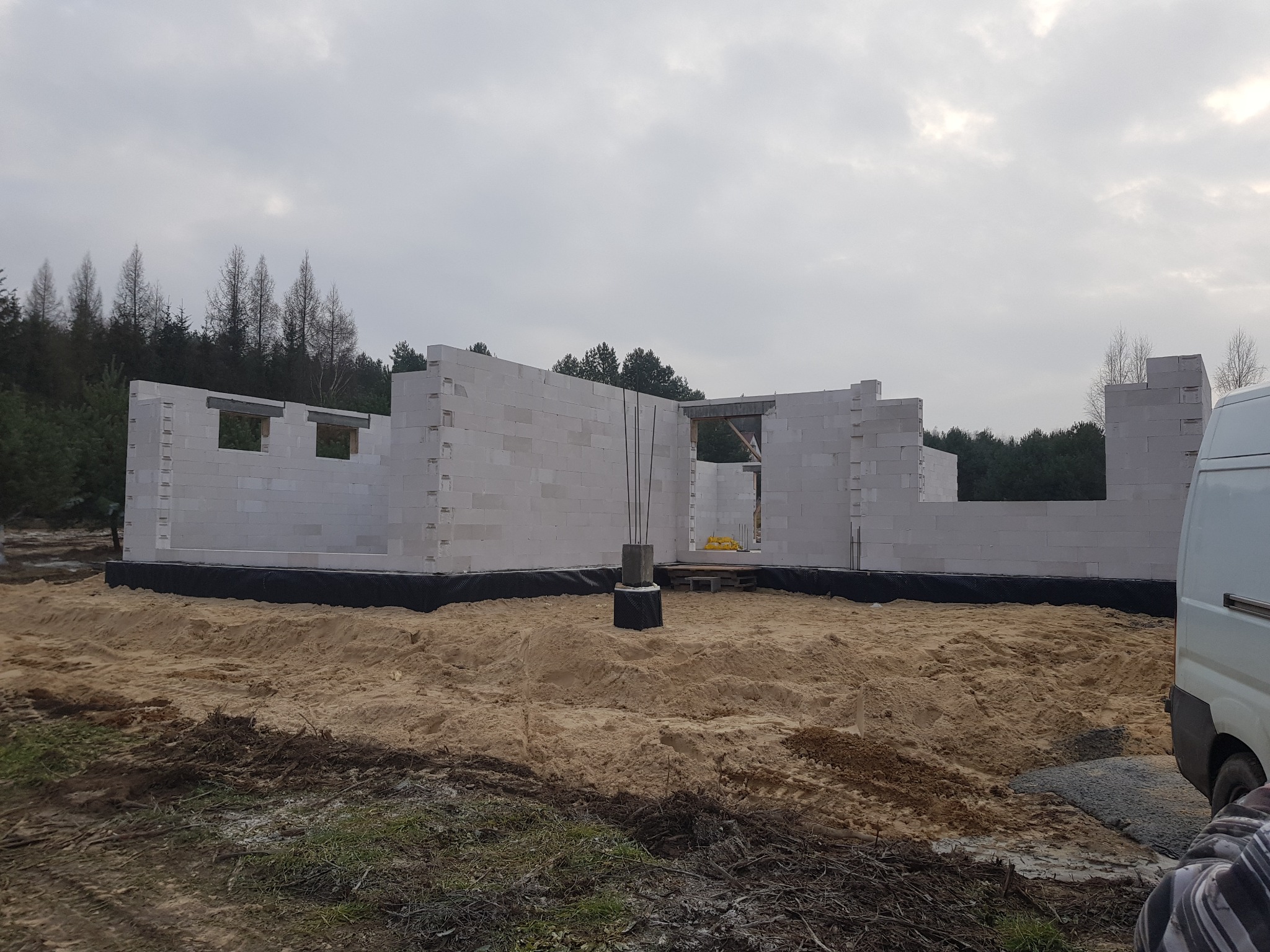 Wznoszone ściany z białych bloczków na etapie budowy domu, widoczne zbrojenia, izolacja fundamentów i piaszczyste podłoże, fragment białego samochodu dostawczego z prawej strony kadru.