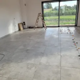 Jasnoszare, kwadratowe płytki podłogowe w trakcie układania, z widocznymi krzyżykami dystansowymi, drabina oparta o ścianę z dużym oknem, widoczne narzędzia i przewody elektryczne przy ścianie.