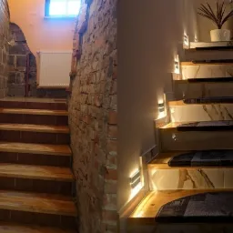 Porównanie schodów przed i po remoncie: surowe cegły i niedokończona ściana po lewej, marmurowe wykończenie z oświetleniem i dywanikami po prawej.