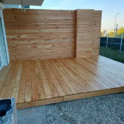 Nowo wybudowany taras z drewnianą ścianką, widoczne świeże drewno i kamienisty grunt wokół.