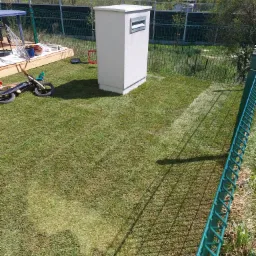 Świeżo założony trawnik z widocznymi śladami po walcu, z boku ogrodzenie z siatki, w tle zabudowa, a z lewej strony fragment drewnianego tarasu z akcesoriami ogrodniczymi i dziecięcy rowerek biegowy.