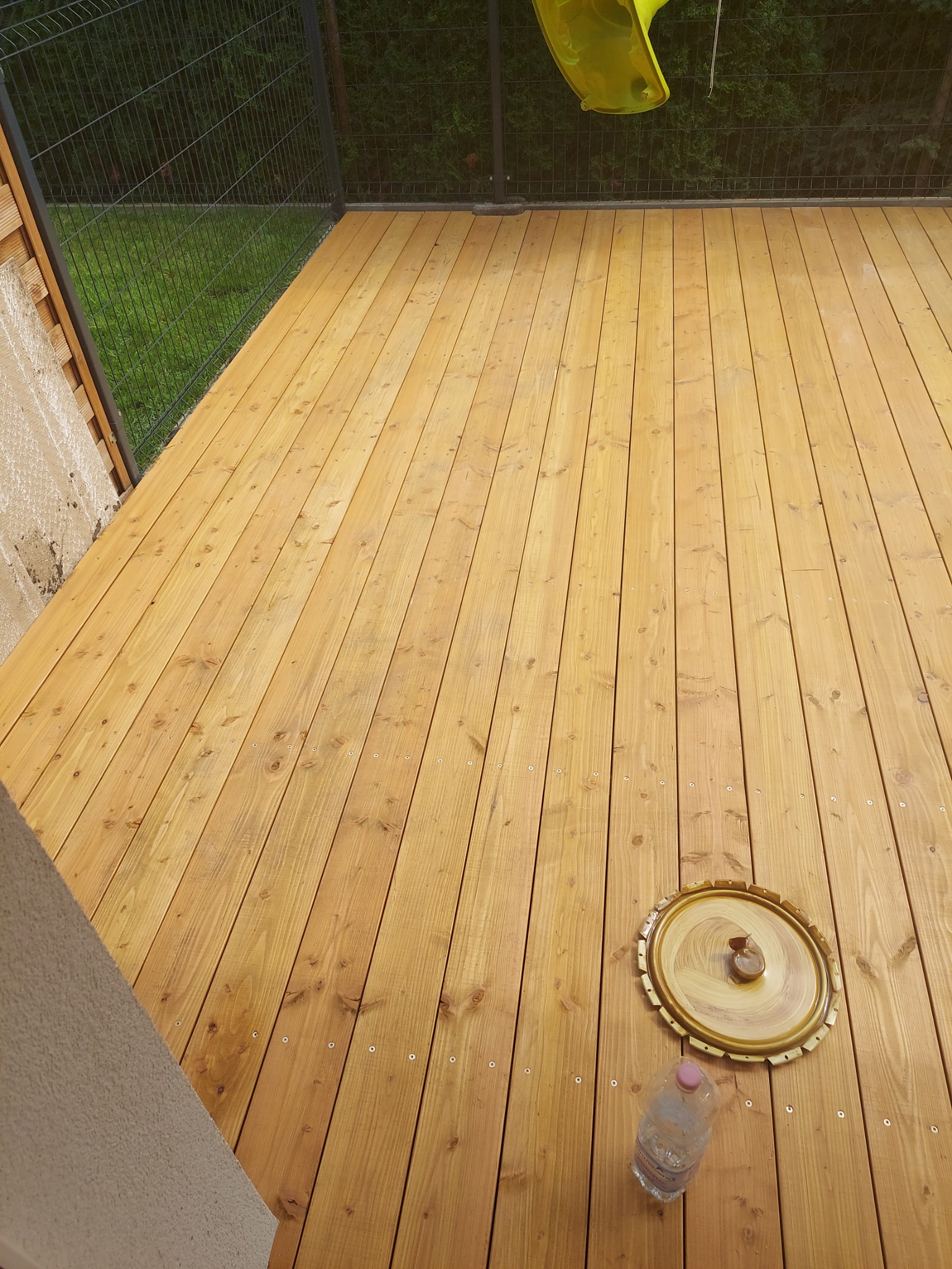 Nowy, jasny taras z desek drewnianych, widok z góry, z fragmentem zjeżdżalni w tle, okrągłą tacą i butelką wody na powierzchni.