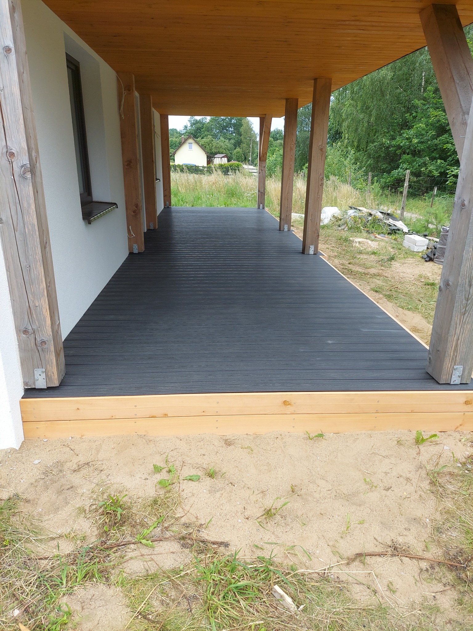 Zadaszony taras z desek kompozytowych w kolorze grafitowym, drewniane słupy nośne, widok na ogród i dom w tle.