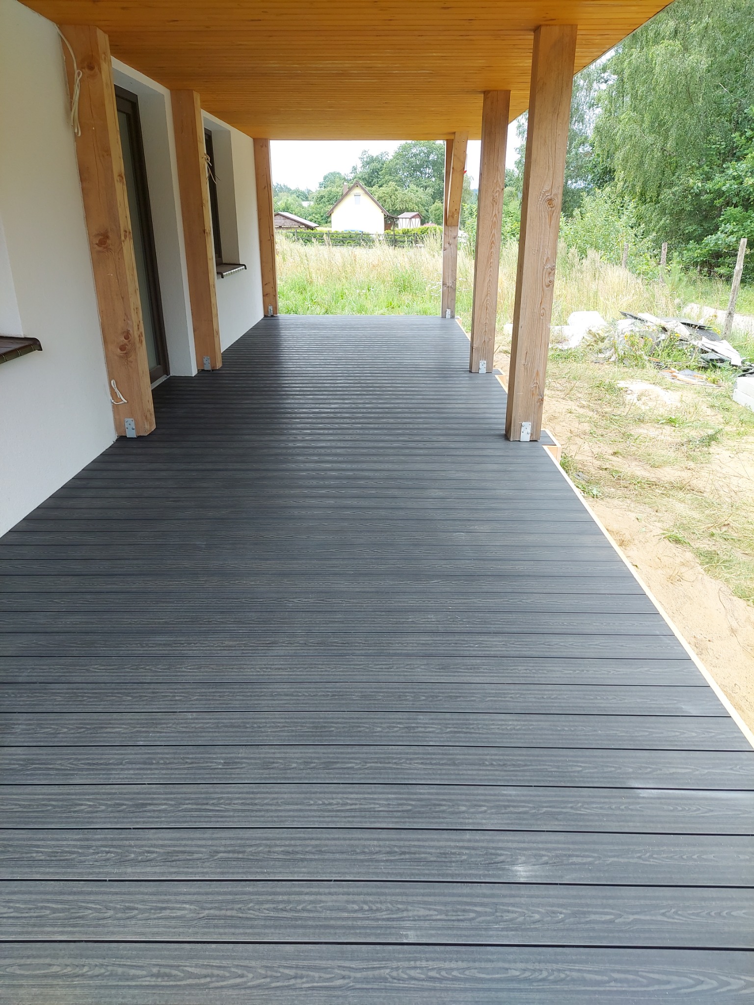 Nowoczesny taras z desek kompozytowych w kolorze grafitowym, z drewnianymi słupami konstrukcyjnymi i widokiem na ogród.