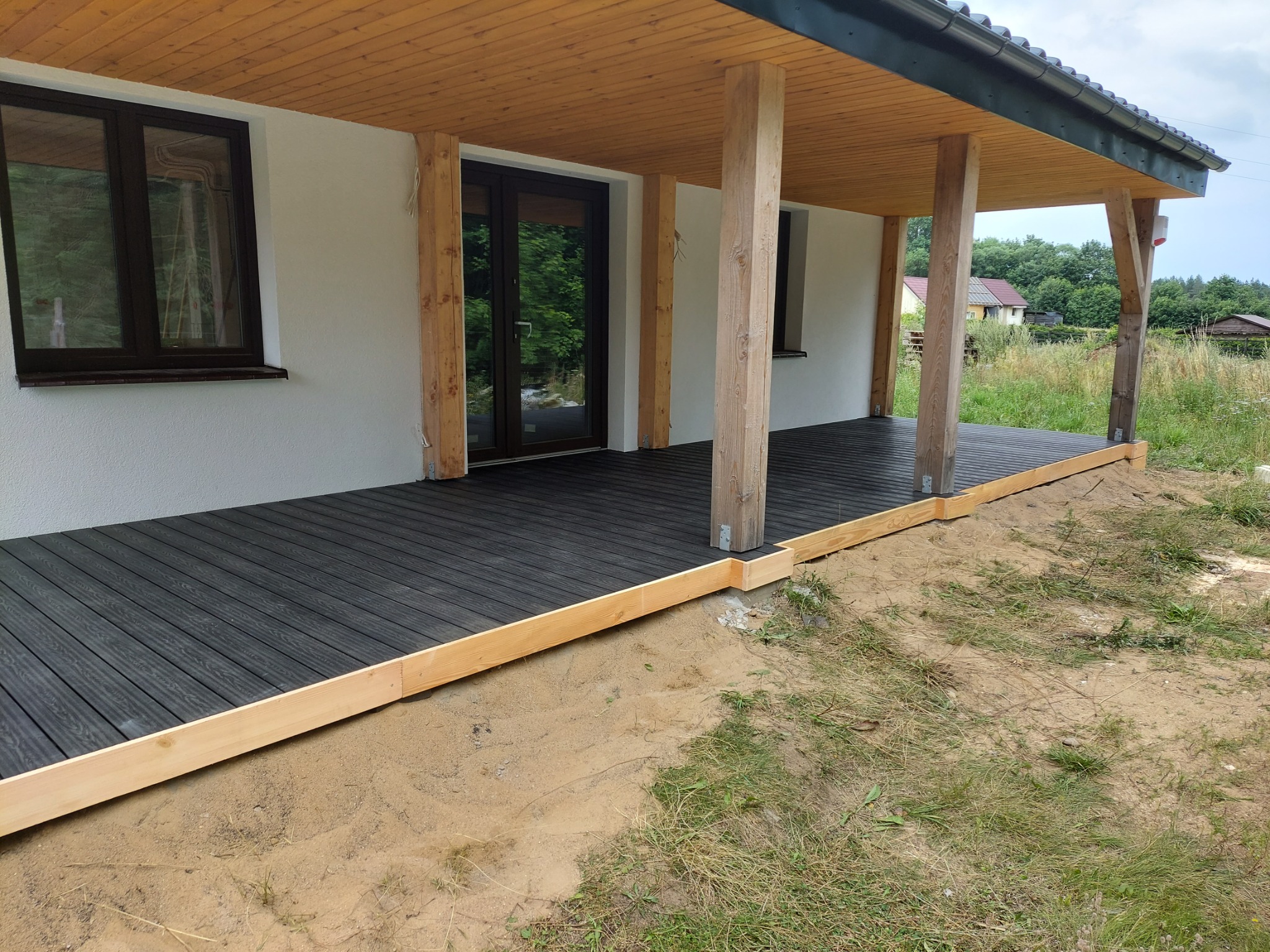 Nowo wybudowany taras z desek kompozytowych w kolorze grafitowym, z drewnianą konstrukcją nośną i słupami, przylegający do białego budynku z ciemnymi oknami i drzwiami, widok na zewnątrz na trawnik...