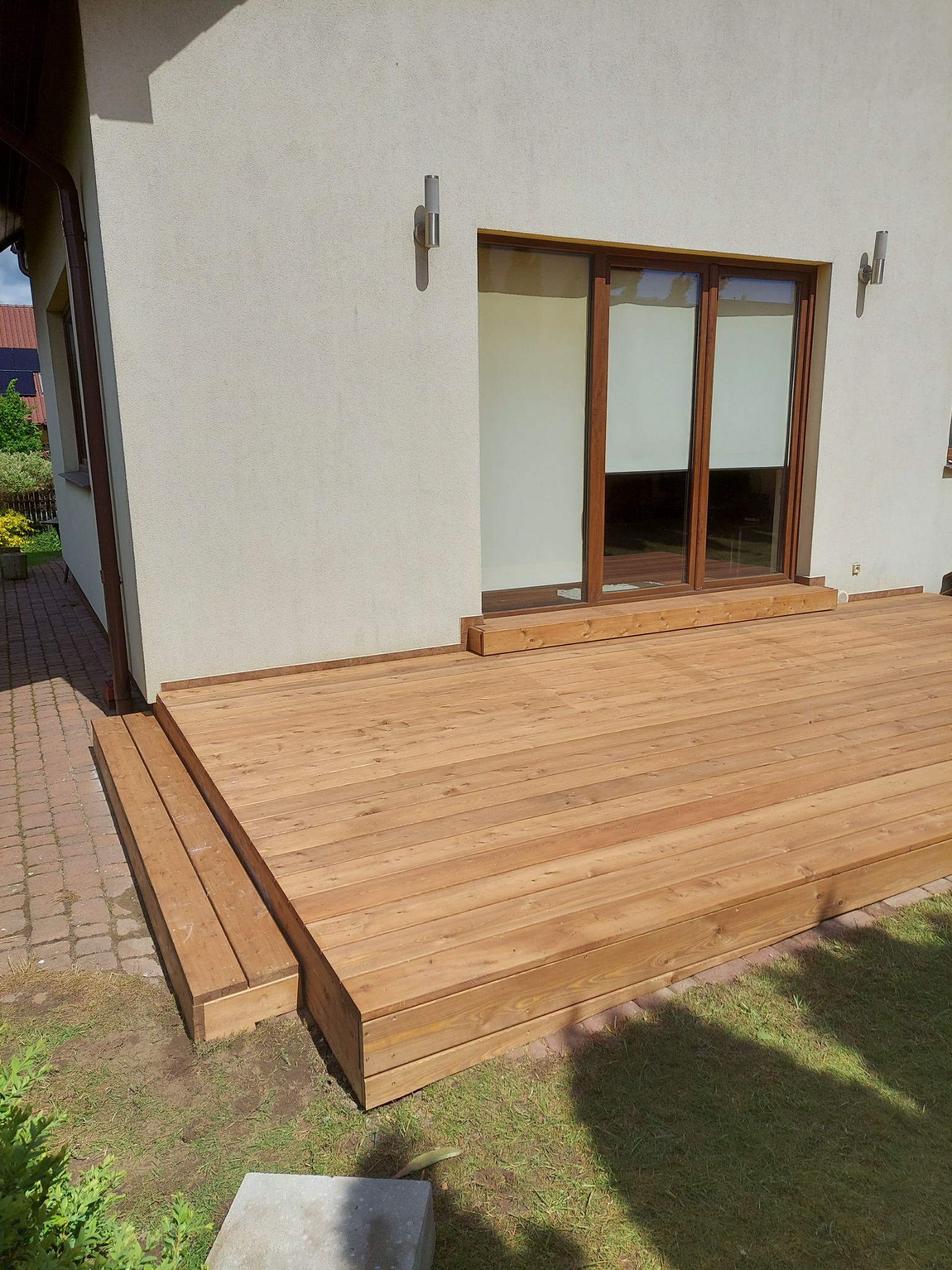 Nowoczesny taras z jasnego drewna przylegający do domu z przesuwnymi drzwiami balkonowymi i zewnętrznymi lampami ściennymi.