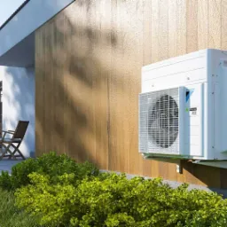 Biała jednostka zewnętrzna klimatyzacji zamontowana na elewacji budynku z drewnianą okładziną, widoczna z poziomu trawnika z krzewami.