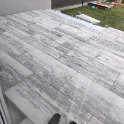 Wyłożony płytami taras z szarym wzorem imitującym drewno, widoczne buty i fragment trawnika z piaskownicą w tle.