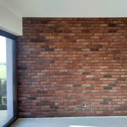 Dekoracyjna ściana z czerwonej cegły w nowym domu, widoczne gniazdka elektryczne i fragment okna z widokiem na zewnątrz.